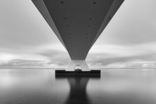 A Dutch coastal landscape in B&W, showing the underside of the Zeelandbrug (Zeeland Bridge) in The Netherlands against a cloudy sky in the back. Under the Bridge - Copyright 2020 Johan Peijnenburg - NiO Photography