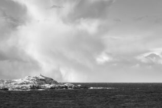 The coast of Lofoten in black & white, featuring a small lighthouse on an island with snow and dramatic storm clouds in the back. Incoming - Copyright 2020 Johan Peijnenburg - NiO Photography