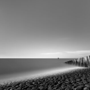 The Dutch coast in Westkapelle, Zeeland, with rocks in the foreground and a breakwater leading the eye to a pole in the North Sea. Captured in black & white. At the Sea - Copyright 2020 Johan Peijnenburg - NiO Photography