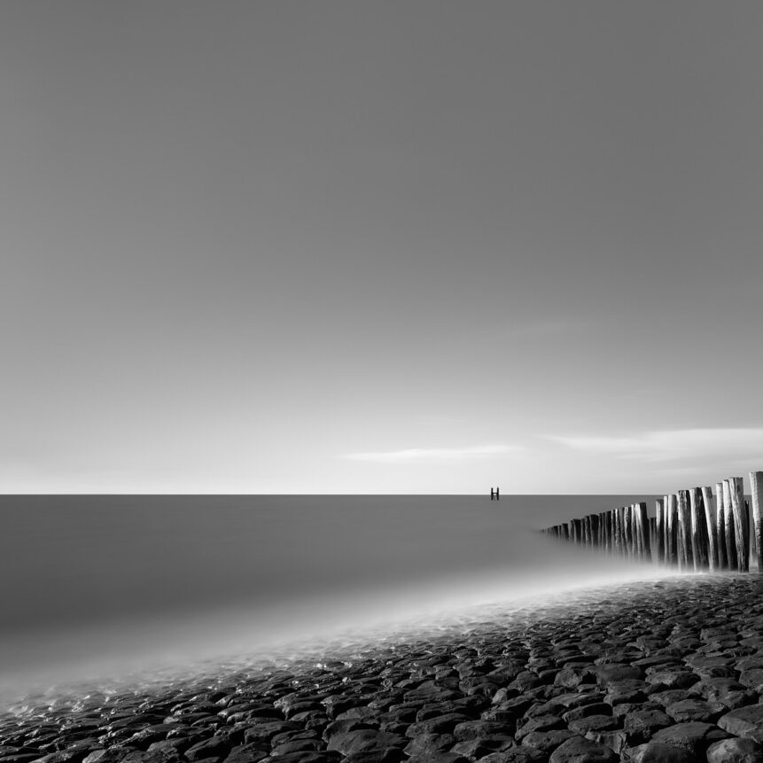 The Dutch coast in Westkapelle, Zeeland, with rocks in the foreground and a breakwater leading the eye to a pole in the North Sea. Captured in black & white. At the Sea - Copyright 2020 Johan Peijnenburg - NiO Photography