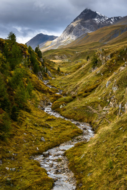 An autumnal landscape, featuring brewing dark clouds above a meadow with a meandering mountain river in the Alps of Graubünden in Switzerland. Up the Stream - Copyright Johan Peijnenburg - NiO Photography