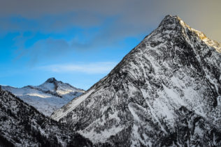 An Alpine winter mountain landscape in colour, featuring two Swiss Alps at sunset with patches of mountain light warming the landscape sideways. Both mountains are located near Saas Fee in Switzerland. Patches of Mountain Light - Copyright Johan Peijnenburg - NiO Photography