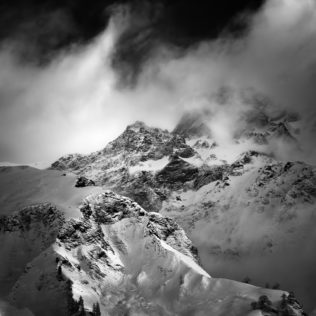 A Swiss mountain landscape in B&W, featuring La Tsavre mountain in winter, surrounded by clouds and fog, near La Fouly in Switzerland. Mystic Mountain - Copyright Johan Peijnenburg - NiO Photography