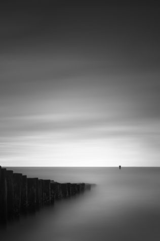 A minimalist Dutch coastal landscape in B&W, with a breakwater in The North Sea leading the eye to a pole and the moody sky at the horizon. Breaking Waves - Copyright Johan Peijnenburg - NiO Photography