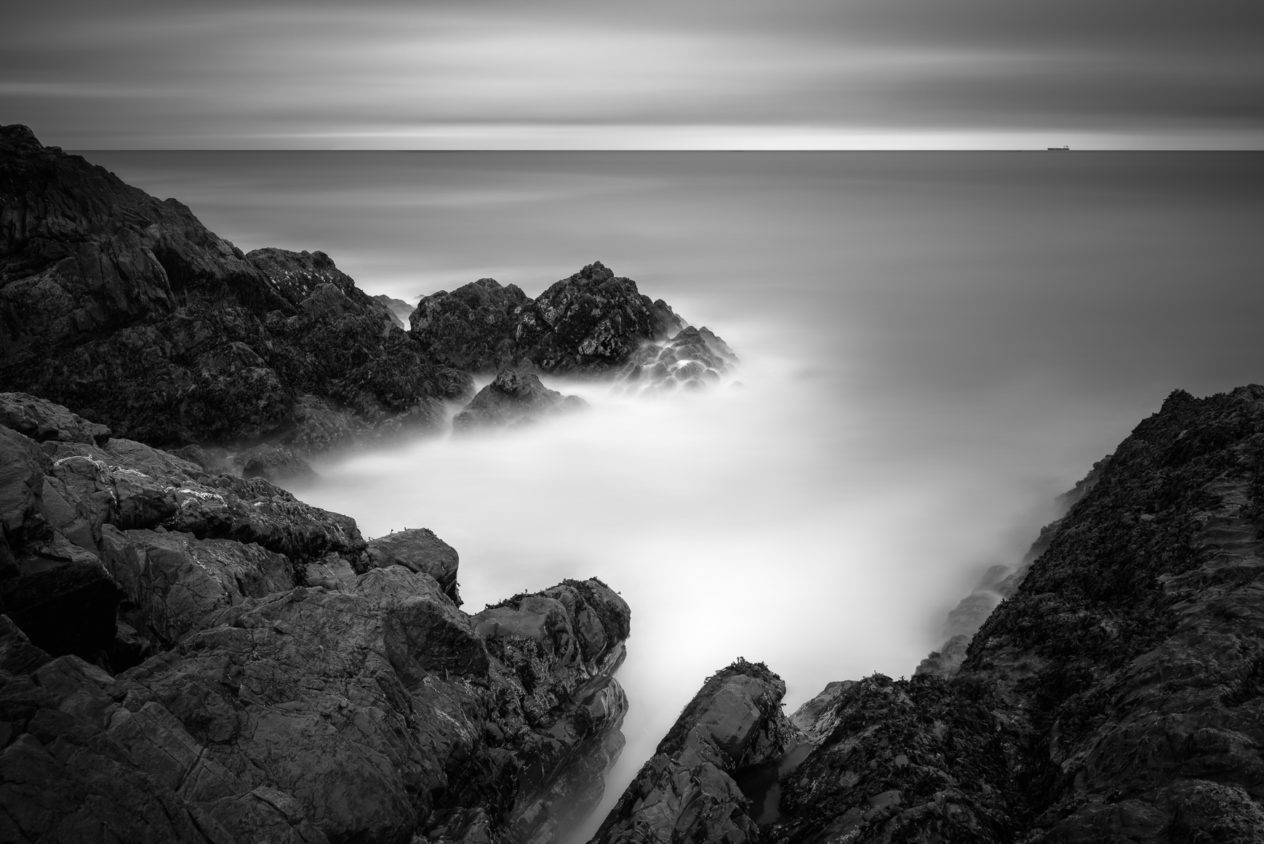 An Irish coastal landscape in black & white showing the rugged cliffs of Greystones Beach and a freight ship in the Irish Sea near Wicklow, Ireland. In the Distance - Copyright Johan Peijnenburg - NiO Photography