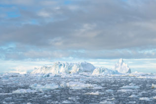 A coastal landscape with icebergs and chunks of ice floating at the shores of Disko Bay near Ilulissat in Arctic Greenland. Soft light warms the icebergs and the clouds sideways. Sea of Ice - Copyright Johan Peijnenburg - NiO Photography