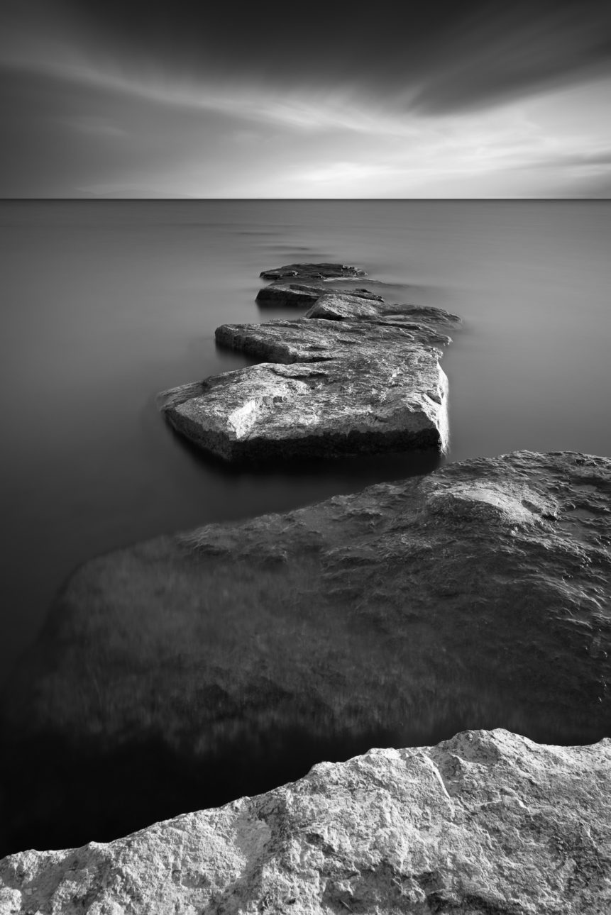 A lake landscape in B&W featuring a line of rocks acting as a breakwater in Lac Léman (Lake Geneva). Walk on Water - Copyright Johan Peijnenburg - NiO Photography