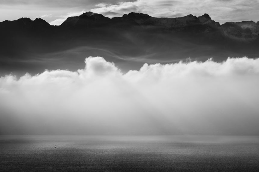 A lake landscape in black & white of Lac Léman and the Alps, with  a sailing boat and the lake lit by sunbeams passing through fog and clouds. Feeling Small - Copyright Johan Peijnenburg - NiO Photography