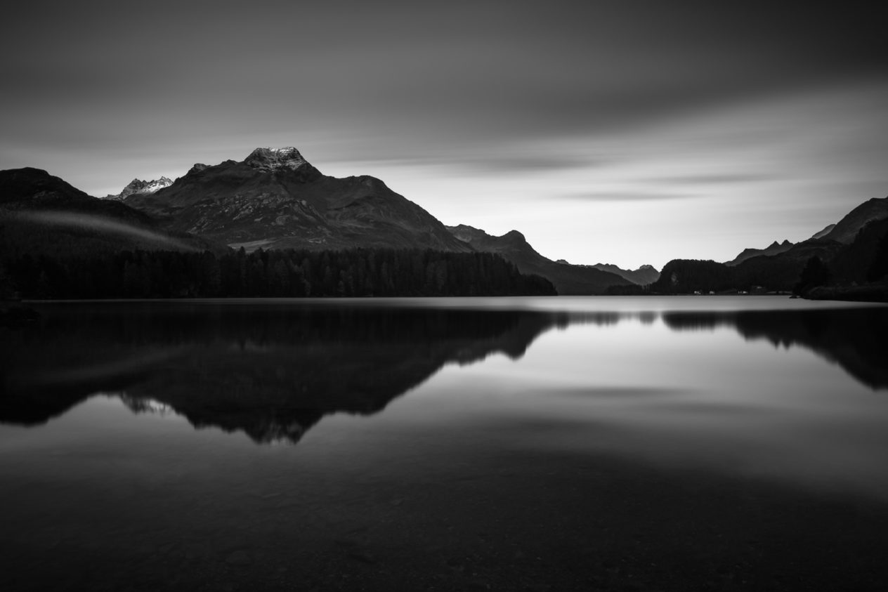 A lake landscape in B&W, featuring the Swiss Alps around Piz de la Margna mountain at sunrise with alpenglow on the peaks and the mountain range reflected in Lake Sils (Silsersee). First Light - Copyright Johan Peijnenburg - NiO Photography