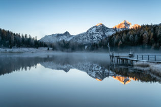 A Swiss mountain lake landscape, featuring a sunrise with alpenglow, a pier and mountain reflections at Lej da Staz (Stazersee) in St. Moritz. A Chilly Rise - Copyright Johan Peijnenburg - NiO Photography