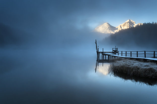 A lake landscape, featuring a misty sunrise at Lej da Staz (Stazersee) with fog, reflections of snow-covered mountains and a wooden pier. Lej da Staz is a small lake in the Swiss Alps near St. Moritz, Switzerland. Misty dawn - Copyright Johan Peijnenburg - NiO Photography