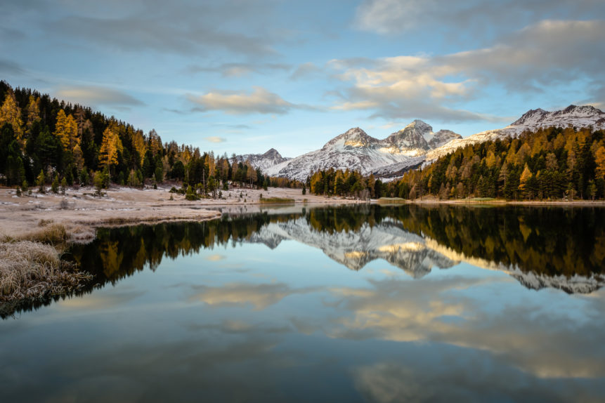 A lake landscape showing a tranquil and colourful sunrise at Lej da Staz (Stazersee) with trees in fall colours and snow on the Swiss Alps. Tranquil Sunrise - Copyright Johan Peijnenburg - NiO Photography