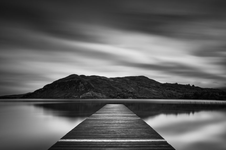 A fine art photograph in black & white, showing a lake landscape with hills and a wooden pier in Lough Caragh in Killarney National Park in Kerry, Ireland. The Landing - Copyright Johan Peijnenburg - NiO Photography