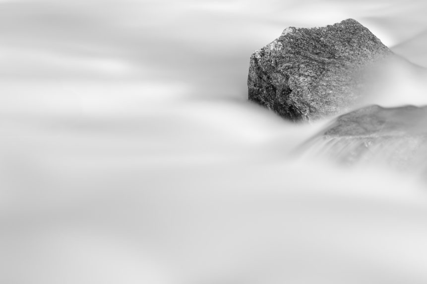 Granite rocks surrounded by the milky water of a glacial mountain river rapid, in a minimalist black and white fine art photograph. Smooth as Silk - Copyright Johan Peijnenburg - NiO Photography