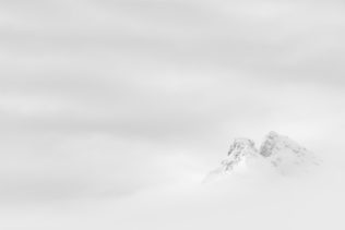 A fine art photograph in black & white, showing a misty mountain landscape with two snow-covered peaks surrounded by thick fog and clouds in Lofoten, Norway. A Glimpse - Copyright Johan Peijnenburg - NiO Photography