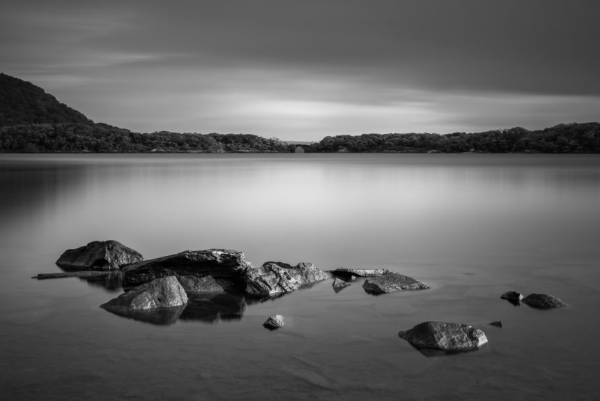 A serene and gentle dawn at Muckross Lake in B&W, with rocks in the foreground and a forest with a stone bridge in the back. Muckross Lake, also called Middle Lake or The Torc, is a lake in Killarney National Park, County Kerry, Ireland. Irish Stillness - Copyright Johan Peijnenburg - NiO Photography