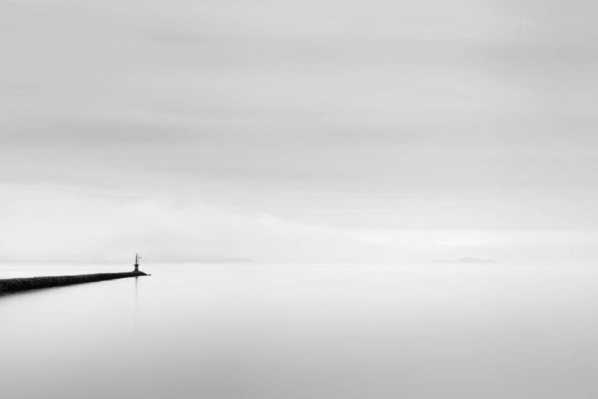 A minimalist lake landscape in black & white, showing a beacon on a jetty in Lac de Neuchâtel in Switzerland on a calm but foggy day. Misty Solitude - Copyright Johan Peijnenburg - NiO Photography