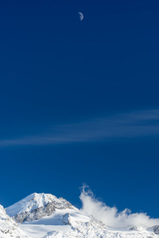 A Swiss winter mountain landscape with snow, with the moon above Piz Tremoggia mountain in Engadin, Switzerland, and fog crawling up in the direction of the moon. Shoot for the Moon - Copyright Johan Peijnenburg - NiO Photography