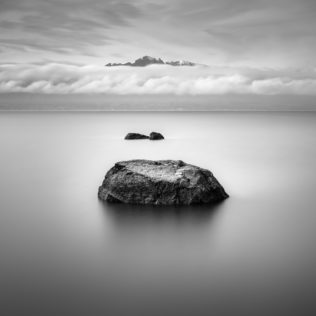 Rocks lined up as stepping stones in Lake Geneva (lac Léman) with the French Alps rising above a band of clouds, in B&W. Stepping Stones - Copyright Johan Peijnenburg - NiO Photography