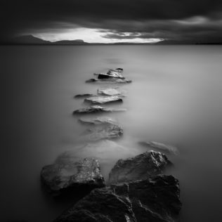 A moody lake landscape in B&W, with a line of rocks in Lac Léman (Lake Geneva) leading the eye to dark clouds and the silhouette of the Alps on the horizon. The End - Copyright Johan Peijnenburg - NiO Photography