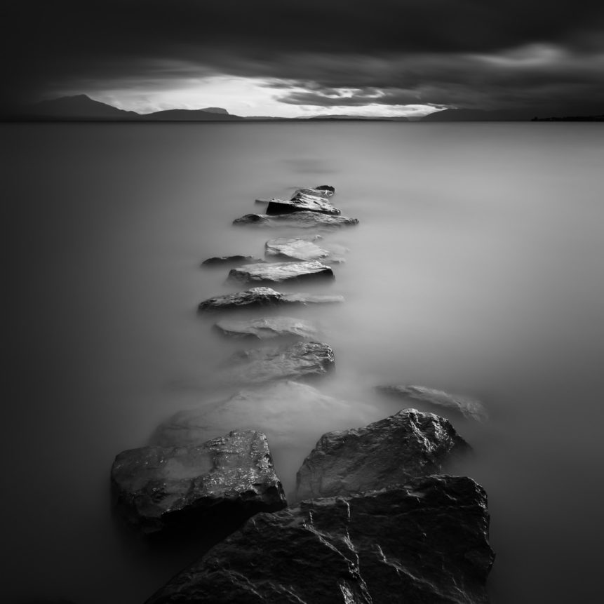 A moody lake landscape in B&W, with a line of rocks in Lac Léman (Lake Geneva) leading the eye to dark clouds and the silhouette of the Alps on the horizon. The End - Copyright Johan Peijnenburg - NiO Photography