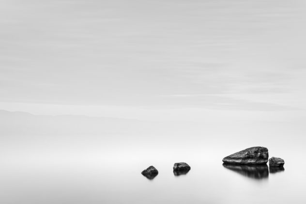 A foggy lake landscape in B&W, featuring four rocks in a quiet mystical Lac de Neuchâtel, with the silhouette of mountains in the back. Stillness - Copyright Johan Peijnenburg - NiO Photography