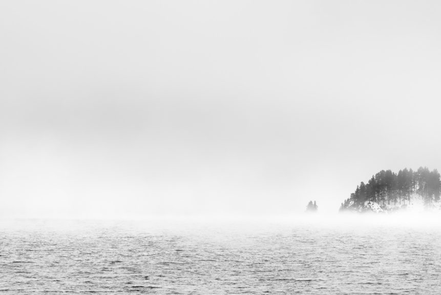 A landscape in black & white, showing a foggy lake landscape at Lake Sils in Engadin with an island with larch trees surrounded by fog. Misty Morning - Copyright Johan Peijnenburg - NiO Photography