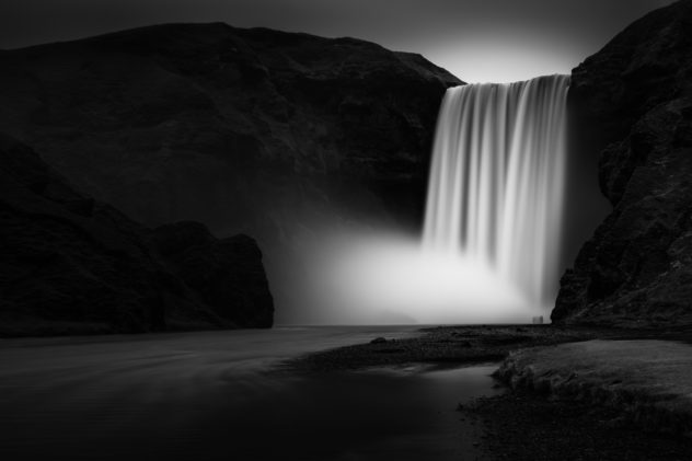 The magnificent Icelandic Skógafoss waterfall in black & white, with people standing in the spray created by the waterfall. Epic - Copyright Johan Peijnenburg - NiO Photography