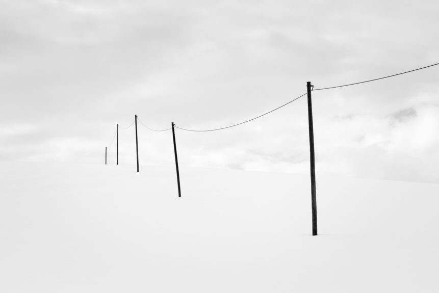 A serene minimalist winter landscape in black & white, featuring a telephony poles in the countryside with snow against a cloudy sky. Out of Sight - Copyright Johan Peijnenburg - NiO Photography