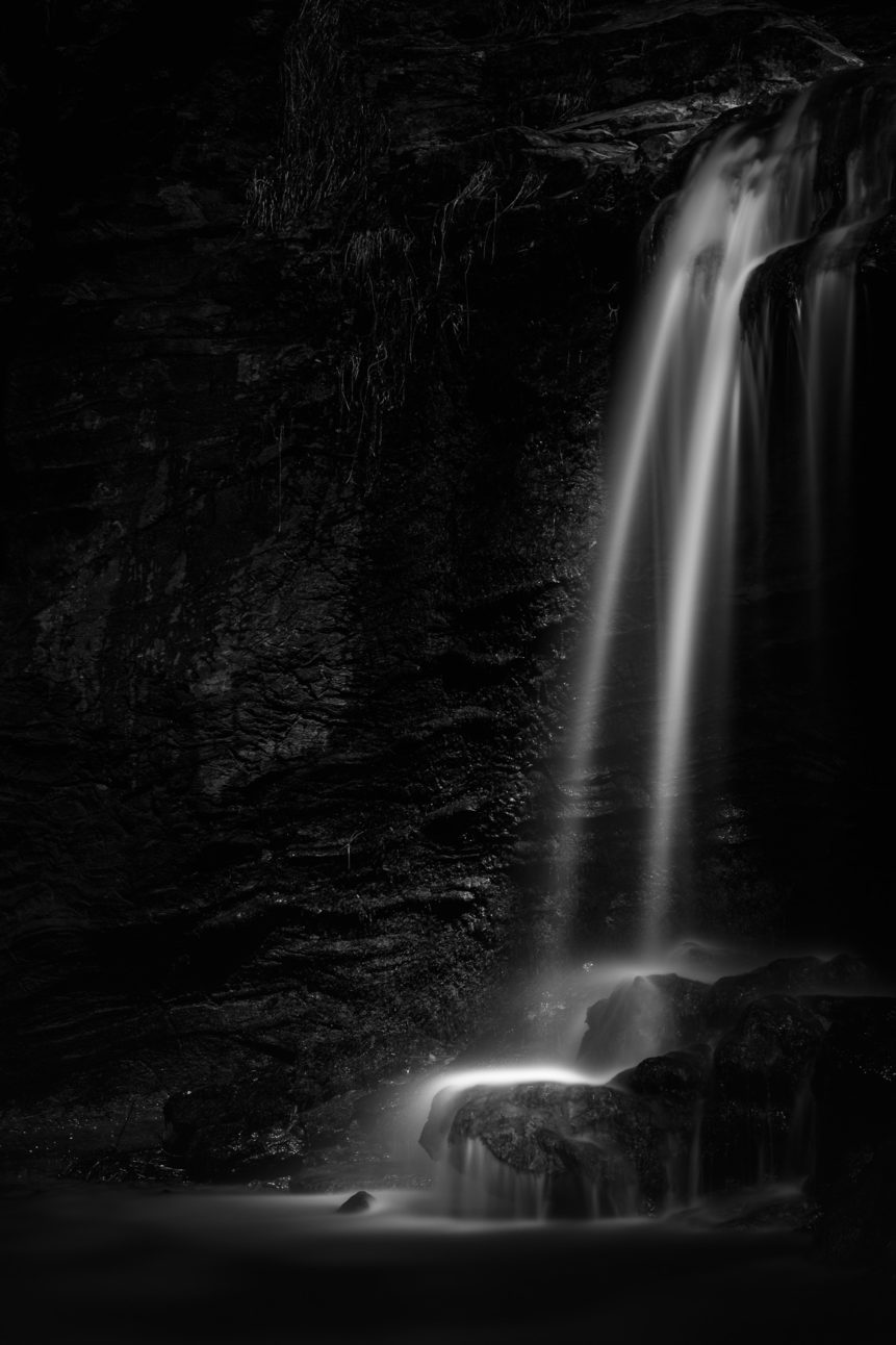 A waterfall in B&W, with water falling from a cliff on rocks. The sun subtly lights the spray of the waterfall. High Falls - Copyright Johan Peijnenburg - NiO Photography