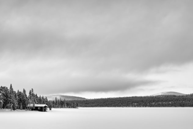 A serene and frosty winter landscape from Swedish Lapland in B&W, featuring a small cabin, a snow-covered lake, a boreal forest, hills, and a big moody sky. Frozen - Copyright Johan Peijnenburg - NiO Photography