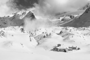 A mountain landscape in black & white, showing a small snowed-in settlement during winter in the Swiss mountains with alpine mountain peaks in the back. Snowed In - Copyright Johan Peijnenburg - NiO Photography