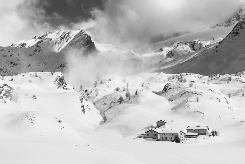 A mountain landscape in black & white, showing a small snowed-in settlement during winter in the Swiss mountains with alpine mountain peaks in the back. Snowed In - Copyright Johan Peijnenburg - NiO Photography